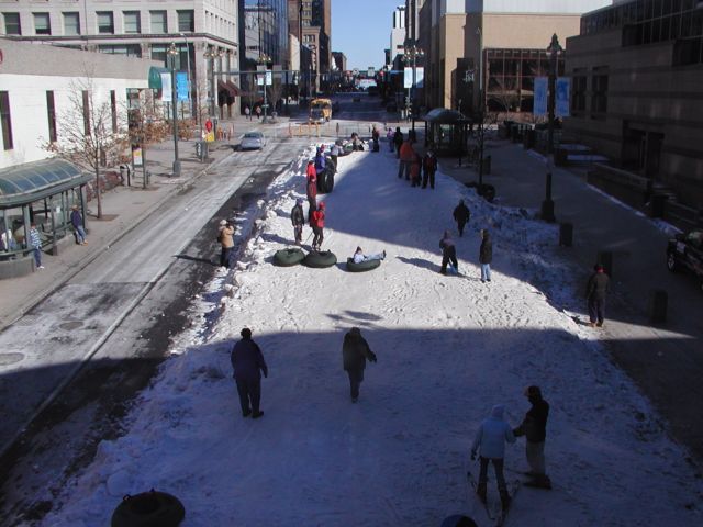 Picture A tube rider soars past those waiting on the sidelines for their turn ride the downtown downhill on East Main St. Rochester NY New York as part of the Coldrush Festival. View looking East on East Main Street at the intersection where Saint Paul St. Becomes South Ave.  February 9th 2003 POD winter Picture Of The Day view picture photo image picture pictures photos images, February 9th 2003 POD