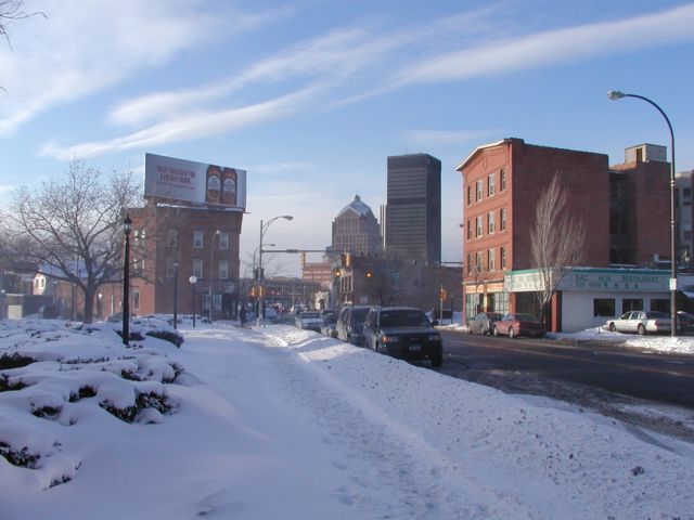 Picture Happy Hour, wind whipped snow blows down a Monroe Ave sidewalk. A billboard perched high above the Bug Jar touts some beer. The Dac Hoa Chinese Barbecue Place is located on the other side of the street. Downtown Rochester NY skyline center background. Rochester NY February 21ST 2003 POD winter Picture Of The Day view picture photo image picture pictures photos images