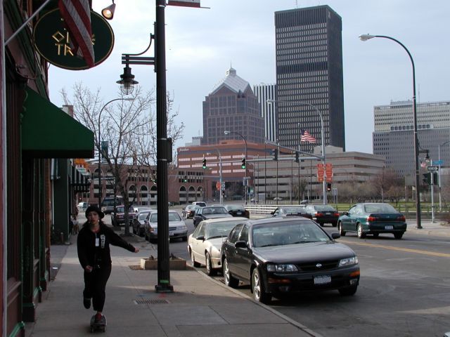 Picture A skateboarder pushes her way past the Olive Tree on Monroe Ave while enjoying a sunny spring afternoon. The downtown Rochester NY skyline fills the background with Frontier Communications, Bausch and Lomb, Lincoln Tower, Xerox, and Midtown Plaza visible.  Picture of the Day Spring I Love NY I luv NY Rochester New York April 19th 2003 POD spring view picture photo image pictures photos images