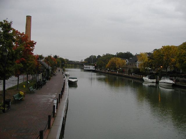 Photo View Looking East from the Lift bridge over the Erie Canal Fairport NY New York, Fall, October 2002, Tree leaves colored green, red, yellow and gold, a few boats tied to both sides of the canal, including a large tour boat, brick paved canal path, a small service bridge in the background. RocPic.Com Rochester NY New York Picture Of The Day 